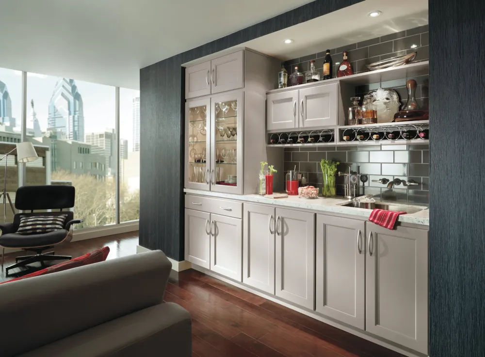 Modern home bar with white cabinets, glass shelves holding bottles and glasses, stainless steel backsplash, and a marble countertop with a sink; large windows show a cityscape outside.