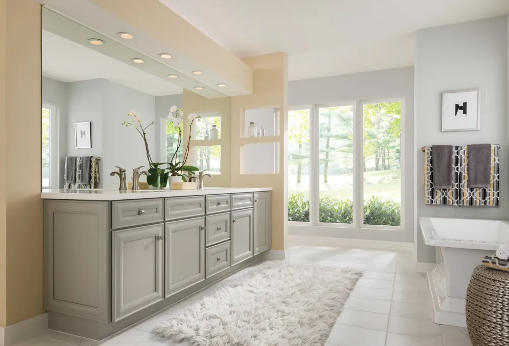 A modern bathroom with gray cabinets, a large mirror, potted plants, a white counter, shelves, a fluffy white rug, and large windows letting in natural light. Towels hang beside a bathtub on the right.