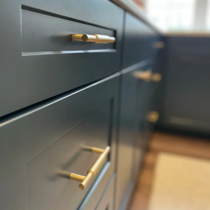 Close-up of dark blue kitchen cabinets with gold horizontal bar handles, shown at an angle. The countertop and a sunlit window are visible in the blurred background.