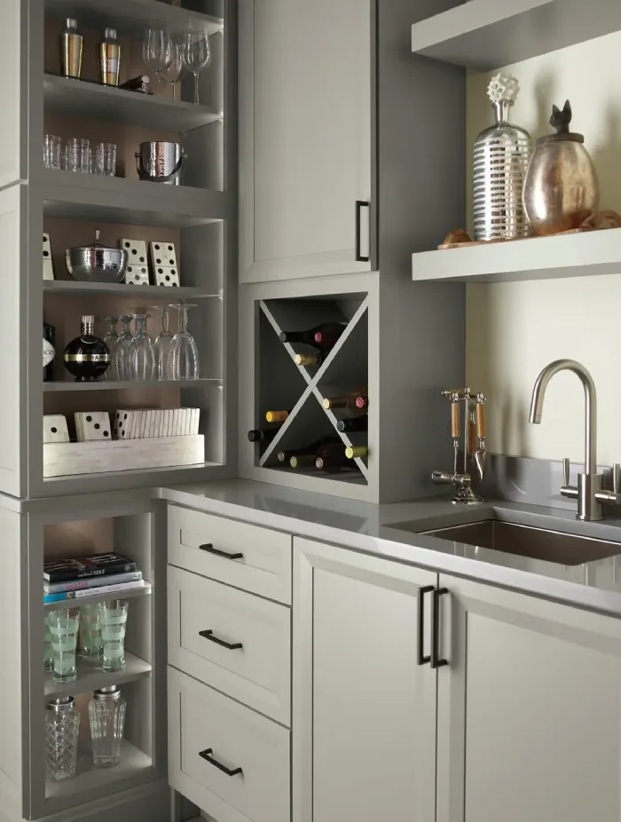 Modern kitchen bar area with open shelves displaying glasses, decanters, a wine rack with bottles, metallic decorative items, and a stainless steel sink with a faucet on a gray countertop.