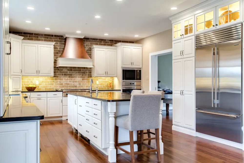 Modern kitchen with white cabinets, stainless steel appliances, a large island with a gray upholstered chair, a brick backsplash, and wooden floors under recessed lighting.