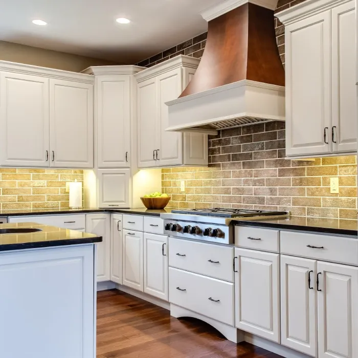 Modern kitchen with white cabinets, black countertops, stainless steel stove, a large copper range hood, brick-style beige backsplash, under-cabinet lighting, and wooden flooring. A bowl of apples sits on the counter.