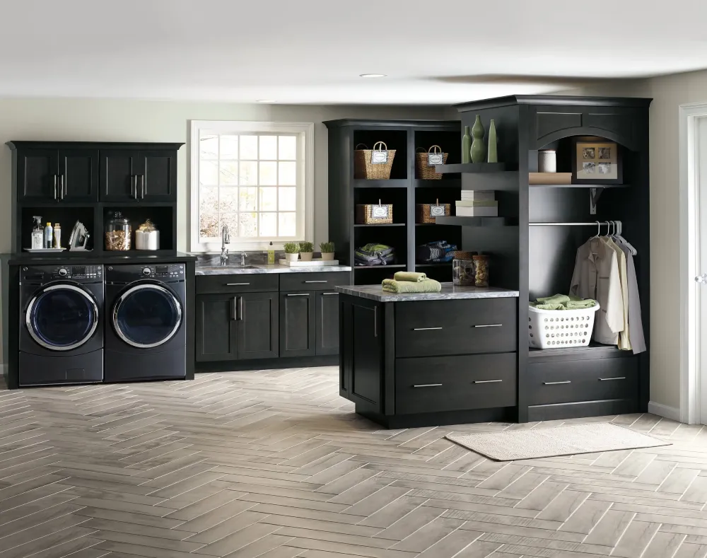 Modern laundry room with dark cabinets, open shelves holding baskets, front-loading washer and dryer, countertop with folded towels, hanging rod, and a large window letting in natural light. Light herringbone tile flooring.