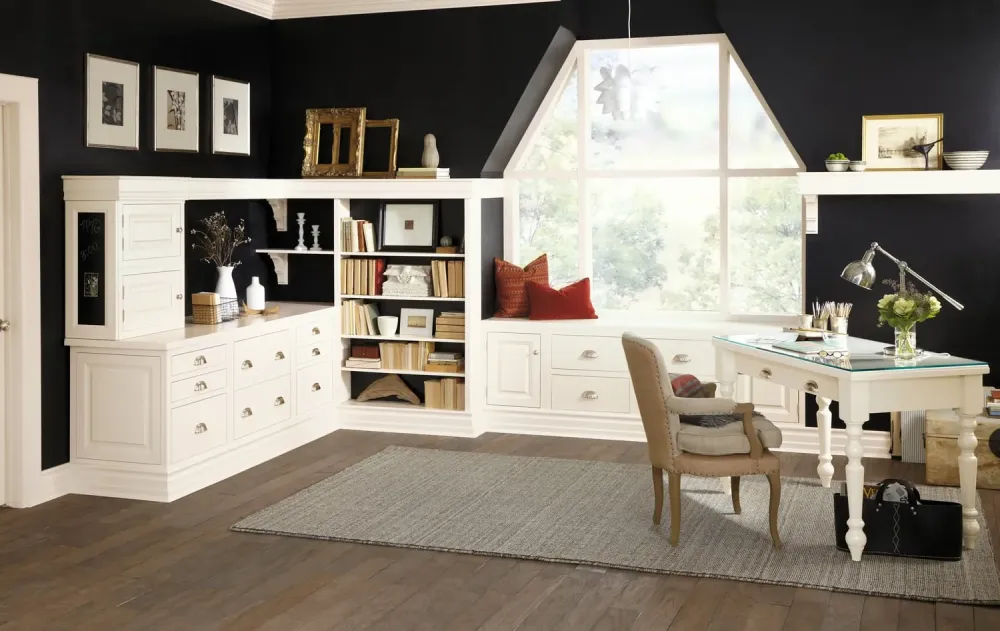 A stylish home office with black walls, white built-in shelves and cabinets, a large window with a seat, a white desk with a lamp and decor, a beige chair, books, and a gray rug on wooden floors.