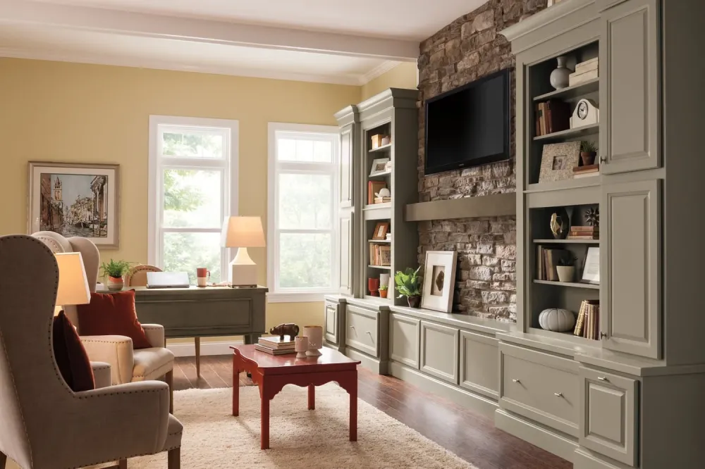 A cozy living room with a stone accent wall, built-in shelves, a mounted TV, neutral-colored furniture, a red coffee table, and large windows letting in natural light. A soft rug covers the wooden floor.