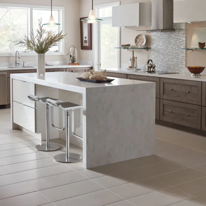 Modern kitchen with a marble island, two metal bar stools, gray cabinets, stainless steel appliances, and a vase with branches. Large windows provide natural light and the backsplash features light gray tiles.