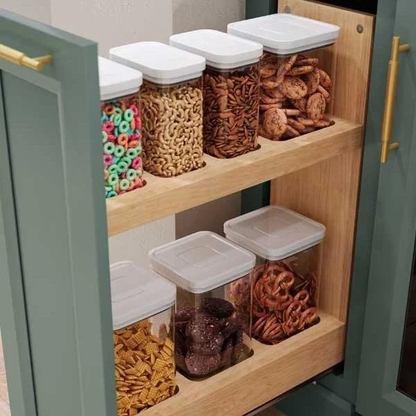 Clear plastic containers with white lids, filled with various snacks like cereal, cookies, pretzels, and crackers, are neatly organized on two wooden shelves inside a green pantry cabinet.