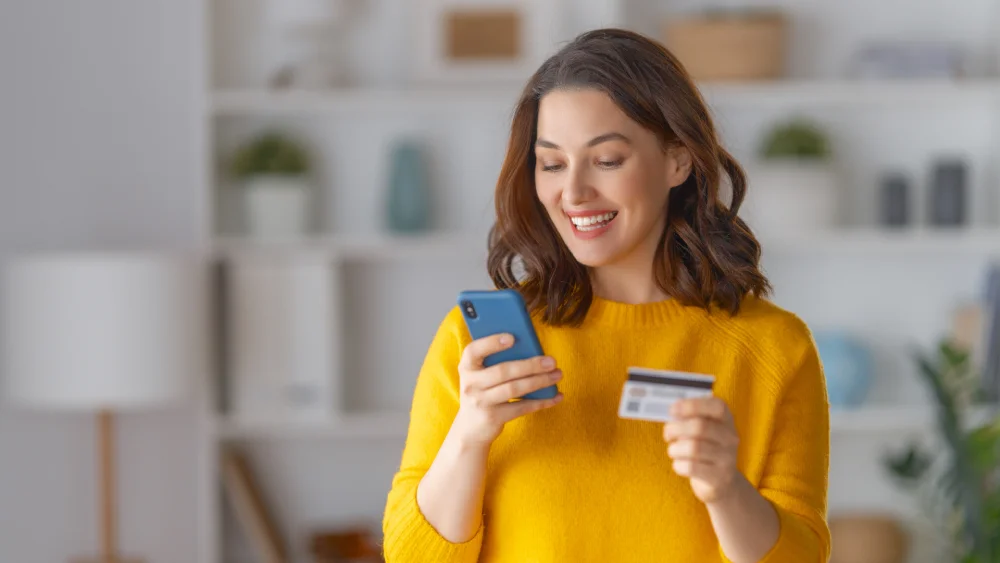 Smiling woman standing in a bright interior while holding a smartphone and credit card.