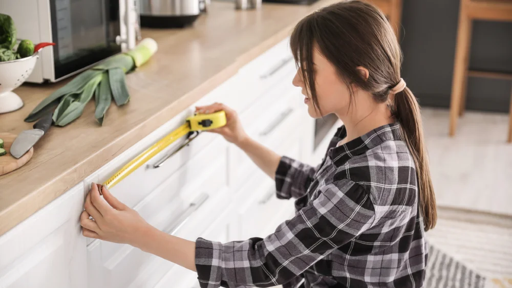 Side view of a female installer using a tape measure along white kitchen base cabinets.