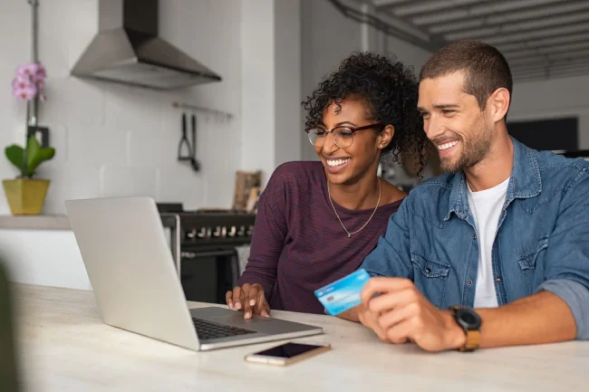 Smiling man and woman sitting together at a table while looking at a laptop computer.
