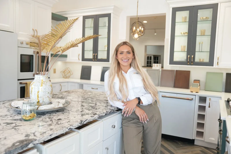 A woman with long blonde hair, wearing a white shirt and khaki pants, stands smiling in a modern kitchen showroom with a marble island, decorative vase, and cabinet samples in the background.