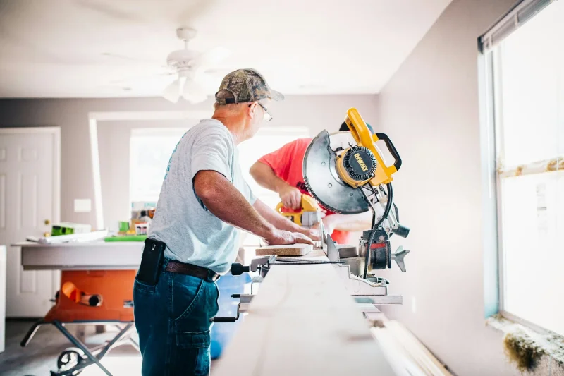 Two construction workers using a power miter saw to cut wood trim indoors.